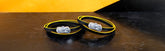Two black and yellow wristbands with transparent clasps rest on a dark, reflective surface against a yellow and black geometric background.