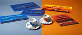 A Lavazza espresso gift set featuring two black Lamborghini-branded cups, matching saucers, a black geometric-patterned plate, and a Lavazza espresso box, all arranged on a dark background.