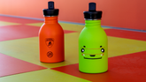 Five camouflage-patterned water bottles and tumblers are displayed on black rocks, surrounded by sand, against a brown background. The items have green, brown, and beige camouflage designs. | Lamborghini Store | Official Website
