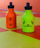 Five camouflage-patterned water bottles and tumblers are displayed on black rocks, surrounded by sand, against a brown background. The items have green, brown, and beige camouflage designs. | Lamborghini Store | Official Website