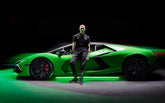 Squadra Corseのアパレルを着た男性が、暗い環境でネオンライトに照らされたグリーンのランボルギーニの隣に立っている。 | Lamborghini Store | Official Website