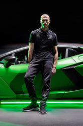 Squadra Corseのアパレルを着た男性が、暗い環境でネオンライトに照らされたグリーンのランボルギーニの隣に立っている。 | Lamborghini Store | Official Website