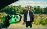 A man stands in a vineyard with rolling green hills in the background, wearing a black jacket and a white t-shirt. A car's side mirror is visible in the foreground, reflecting greenery. | Lamborghini Store | Official Website