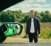A man stands in a vineyard with rolling green hills in the background, wearing a black jacket and a white t-shirt. A car's side mirror is visible in the foreground, reflecting greenery. | Lamborghini Store | Official Website